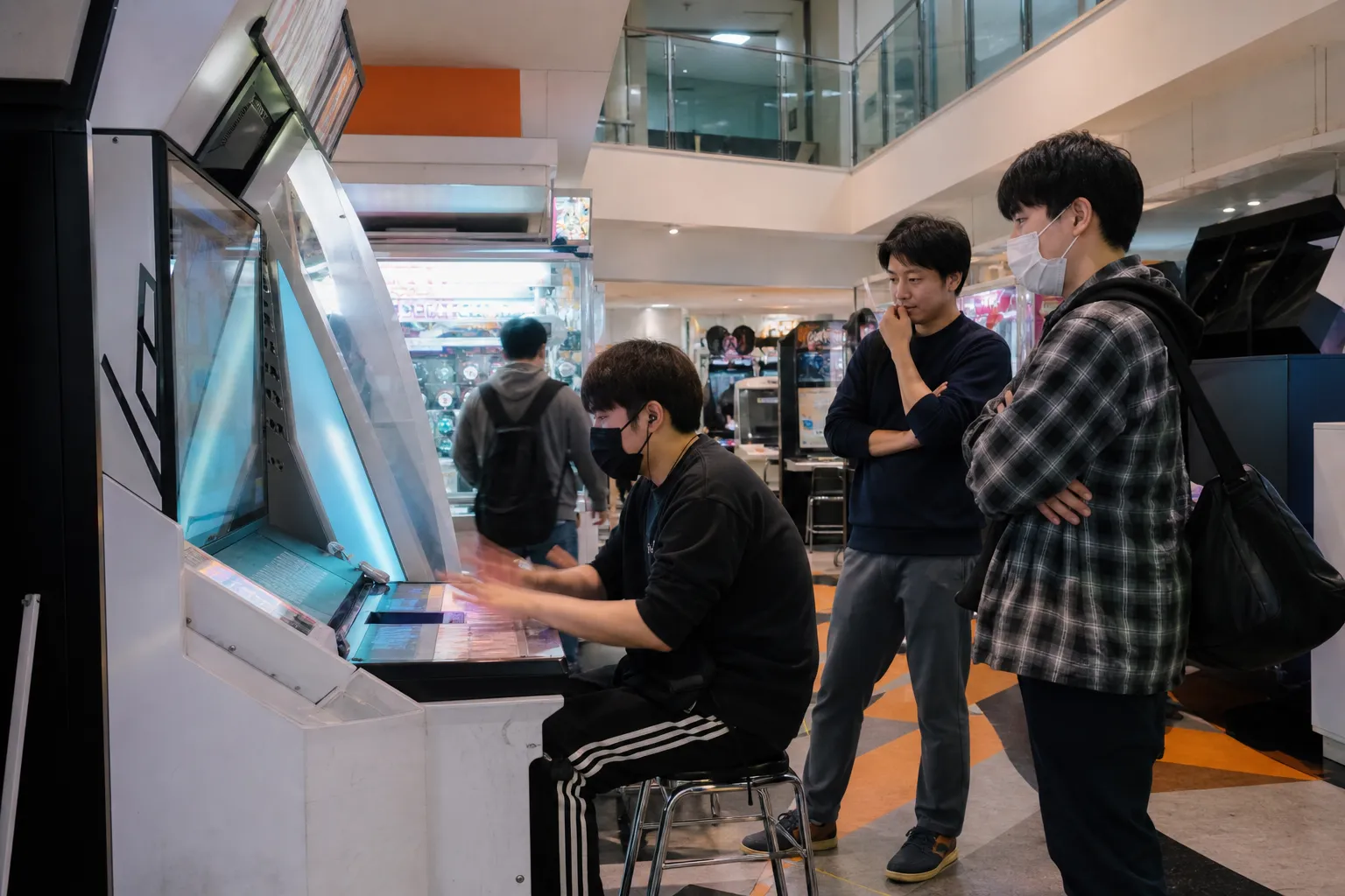 日本のゲームセンターで、音楽ゲームに集中する人を二人の来店者が少し離れて見守る様子。