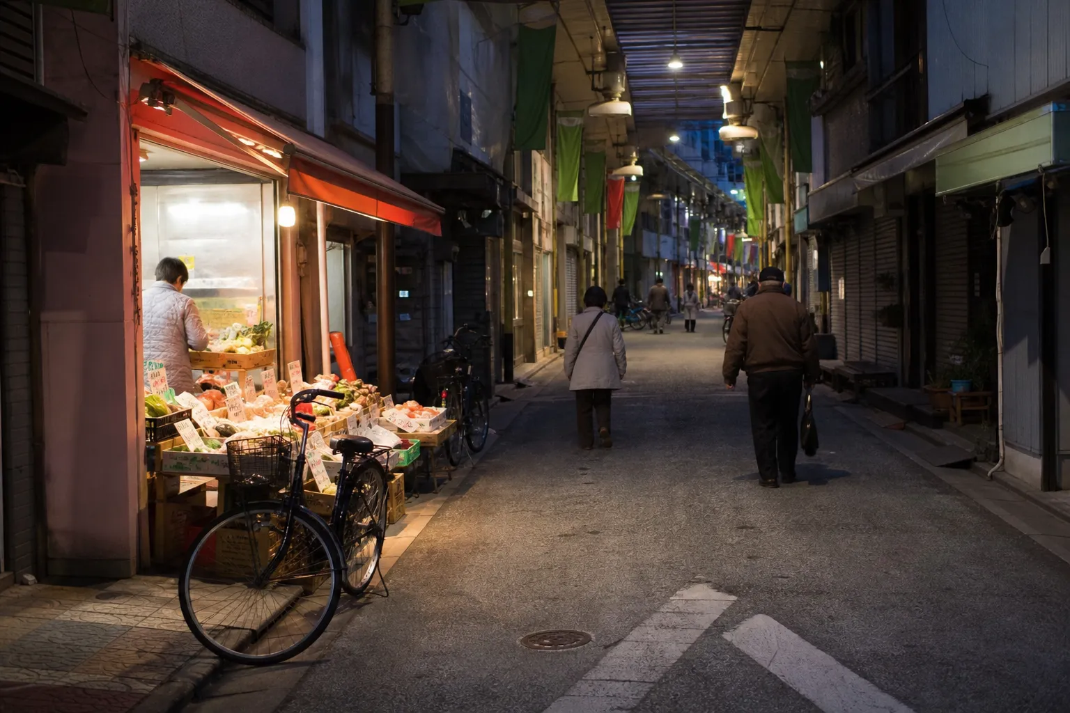 夕暮れの地域商店街で、青果店の灯りのそばを住民が歩く様子。