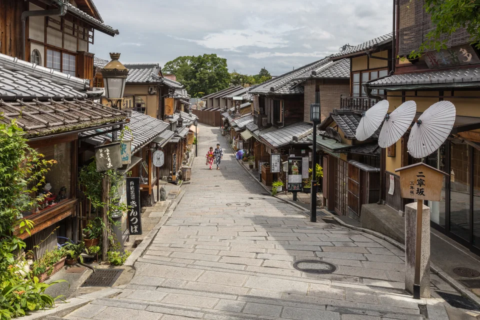 People walking along the stone-paved Ninenzaka street, with wooden shopfronts and paper umbrellas in Kyoto.